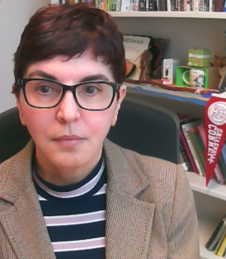 A woman with short dark hair and glasses in front of a book case with a Cornell pennant